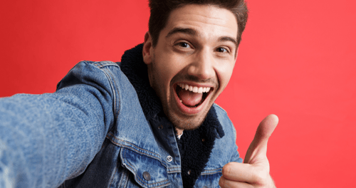 Iloinen ja hymyilevä mies denim-takissa ottaa selfietä ja näyttää peukaloa ylöspäin punaisella taustalla kuvastaen tyytyväistä kanta-asiakasta.
