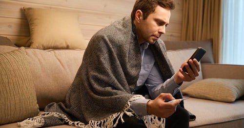 Vilttiin kääriytynyt mies istuu sohvalla katsoen huolestuneena älypuhelintaan kuumemittari kädessään kuvastaen sairastumista ja digitaalisen vakuutuskortin tarvetta.