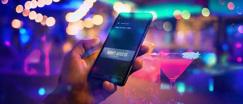 Kädessä oleva älypuhelin, jonka näytöllä näkyy NoHo Partnersin digitaalinen NightAccess Blue -kanta-asiakaskortti värikkäästi valaistussa yökerhoympäristössä cocktail-lasien vieressä.